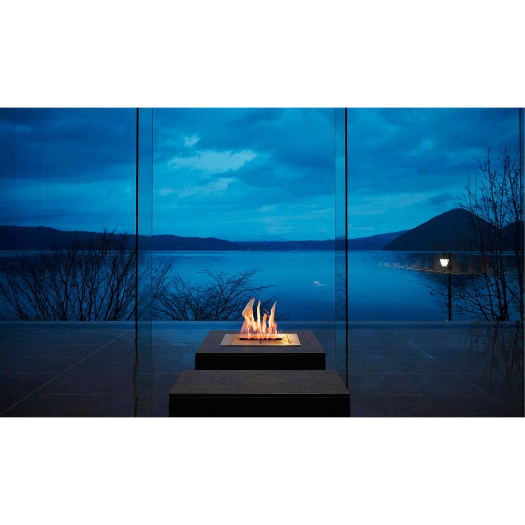 EcoSmart Fire BK5 ethanol burner with a golden flame set in a dark square base before a lake view at dusk.