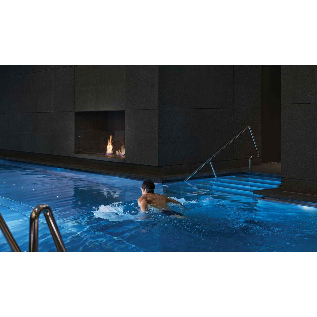 EcoSmart Fire BK5 ethanol burner integrated into a dark tiled wall above a blue-lit indoor swimming pool.